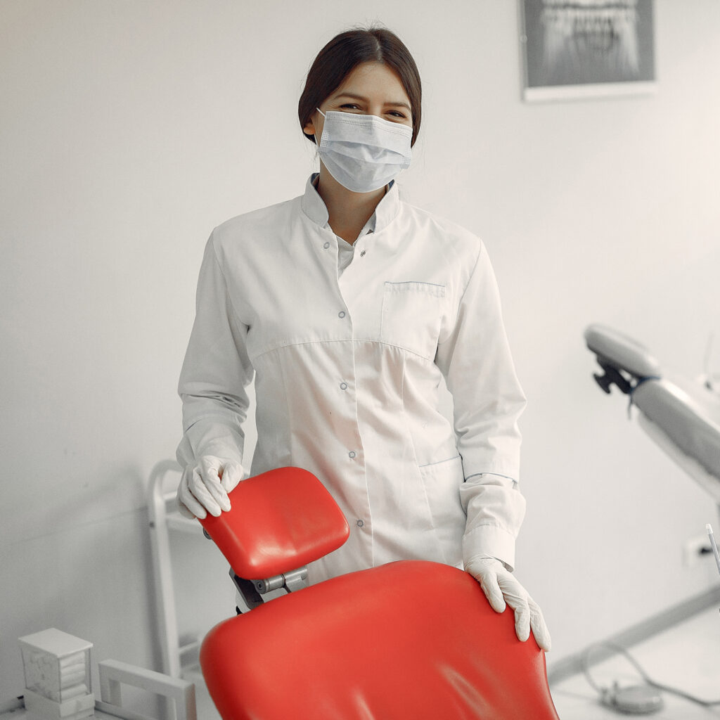 Zahnärztin mit Maske neben rotem Zahnarztstuhl im Behandlungsraum.